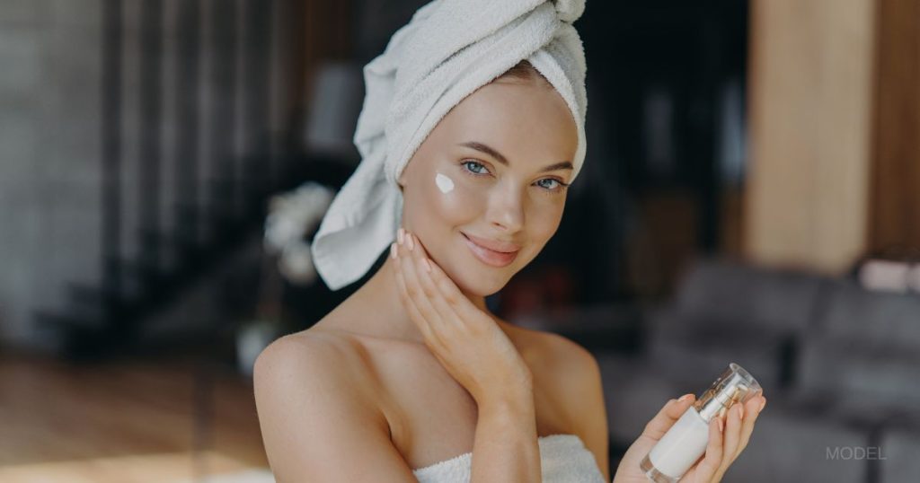 Beautiful woman with a towel around her hair and lotion on her face, holding a skin care bottle. (MODEL)